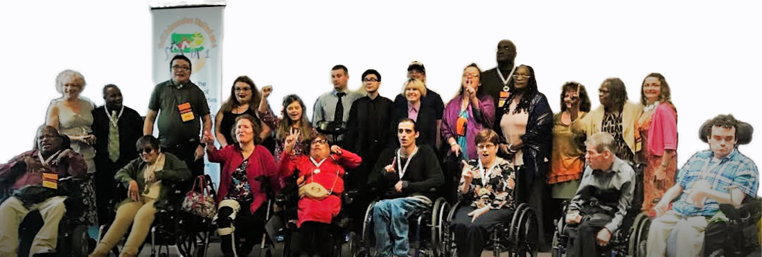 Twenty three people smiling and holding up one finger to show they are united as one. Some of them are standing, some using wheelchairs.